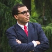 A person wearing glasses and a suit with a red tie stands confidently outdoors, arms crossed, surrounded by lush green trees.