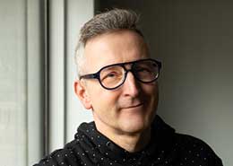 A person with glasses and a black dotted shirt stands by a window, smiling softly. The background is softly lit and neutral.