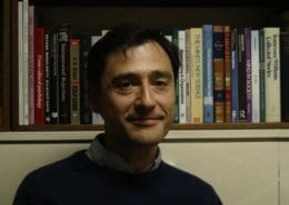 A person stands in front of a bookshelf filled with various books, smiling subtly. The books vary in color and appear scholarly.