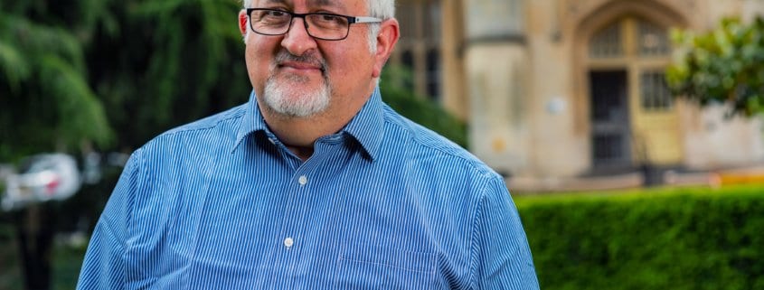 A person with glasses and a striped shirt stands outdoors with a historic building in the background.