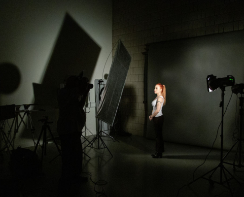 A person poses in a dimly lit photography studio, with equipment and lighting creating intricate shadows on the wall.