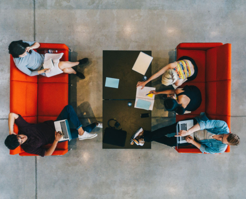 Five people collaborate around a table with laptops and notebooks, sitting on red couches in a modern space with concrete flooring.