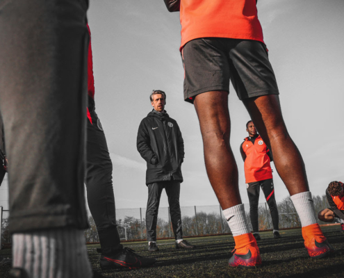 A group of people in sports attire stand on a football field, listening to a person in the center. Overcast sky above.
