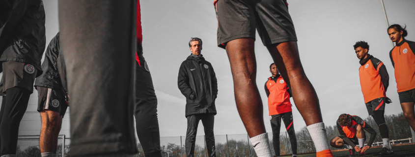 A group of people in sports attire stand on a football field, listening to a person in the center. Overcast sky above.