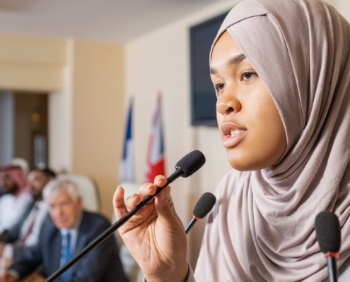 A person speaks at a conference, holding a microphone. International flags are visible in the background, suggesting a multinational event setting.