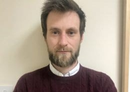 A person with a beard stands against a plain wall indoors, wearing a maroon sweater. No landmarks or historical buildings are visible.