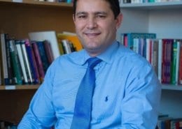 A person in a blue shirt and tie sits smiling in a library, with numerous books visible on shelves behind them.