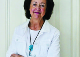 A person wearing a white jacket and a turquoise pendant stands against a light-colored wall, smiling gently with folded arms.
