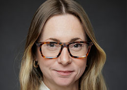 A person with glasses and long, blonde hair is in a portrait against a dark background, wearing a white top with a slight smile.