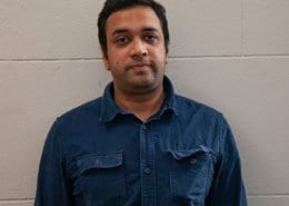 A person wearing a dark blue shirt stands against a plain light-colored wall, looking directly at the camera.