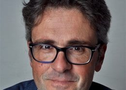 A person with short, curly hair and glasses, wearing a blue jacket. The background is plain and does not feature any landmarks.