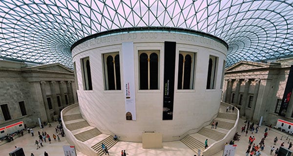 The British Museum's Great Court with its iconic glass ceiling, surrounds the circular Reading Room. People are walking and exploring the vast space.