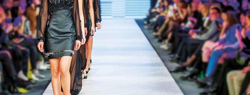 A fashion show scene displays models walking down a runway, surrounded by an audience, highlighting elegant attire in a vibrant, illuminated setting.