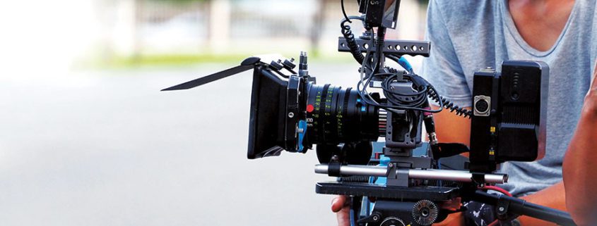 A person operates a professional video camera rig outdoors, focusing intently, with blurred greenery and fencing in the background.