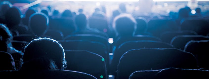An audience sitting in theater seats faces a brightly lit stage, with silhouettes of people visible. Scene suggests a performance or movie.