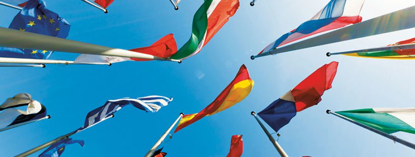 Various national flags, including the European Union, are waving on poles against a clear blue sky, viewed from below, symbolizing international unity.