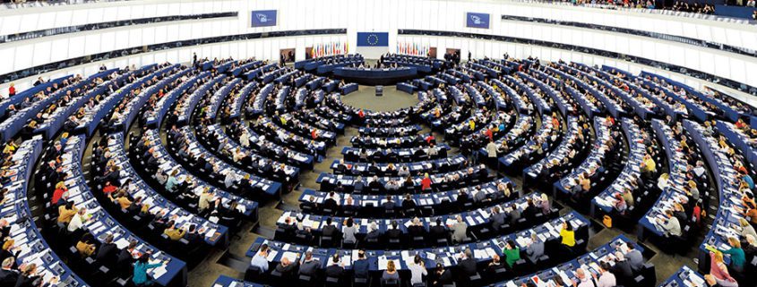 People seated in a large, semicircular auditorium, likely the European Parliament. The room is well-lit with high ceilings and numerous desks.