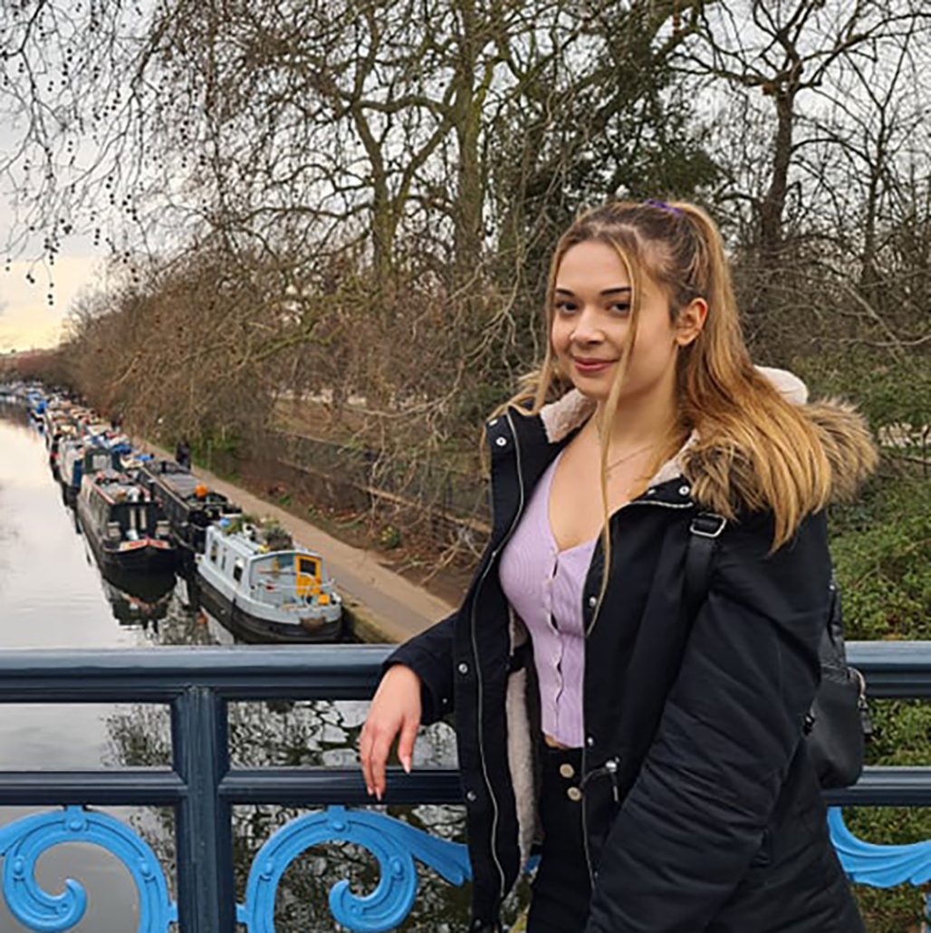 A person stands on a bridge over Regent's Canal, with moored boats and winter trees lining the waterway.