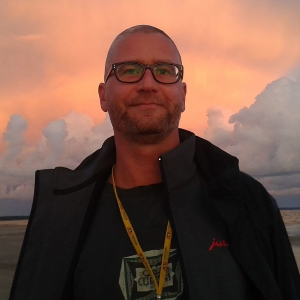 A person with glasses stands outdoors; a vibrant sunset sky with clouds forms the backdrop. The person wears a jacket and lanyard.
