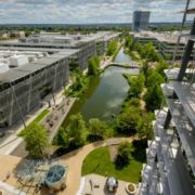 Aerial view of a modern business district with a central waterway, green spaces, and walkways, surrounded by office buildings and a tall structure.