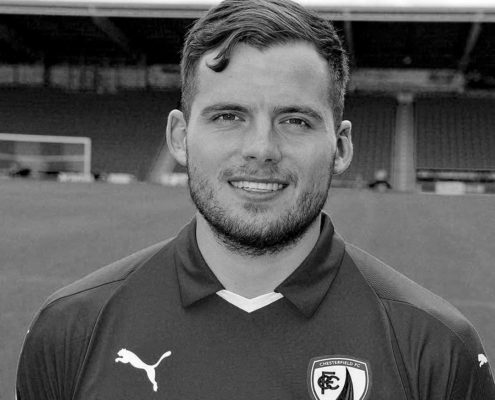 A person in a Chesterfield FC jersey stands in a stadium. The image is in black and white, showing the person smiling.