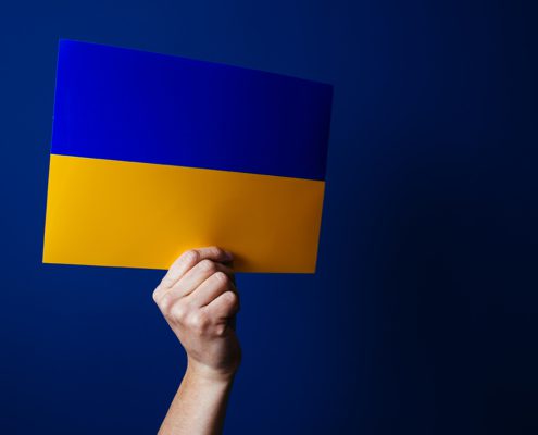 A person holds the blue and yellow Ukrainian flag against a solid blue background, symbolizing support and national pride.