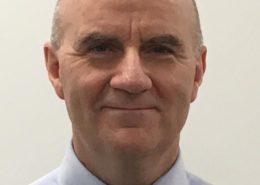 A person with a bald head is wearing a light blue shirt and a dark tie, against a plain white background.