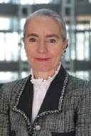 A person wearing a blazer suit, a necklace, and a fabric collar is smiling indoors with their chin tilted up.