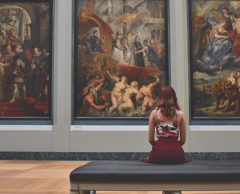 A person in an art gallery sits, viewing large classical paintings depicting historical and mythological scenes on the wall.