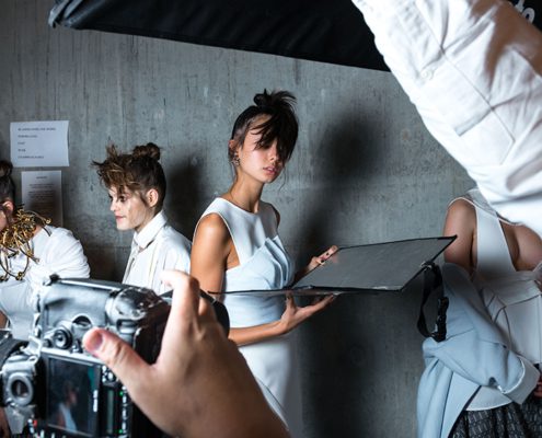 Fashion shoot setting with several people in stylish outfits being photographed. The atmosphere is professional, with lighting equipment and a concrete backdrop.