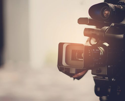 A person operates a large professional video camera, focusing intently. The background is blurred, emphasizing the equipment and the person's action.