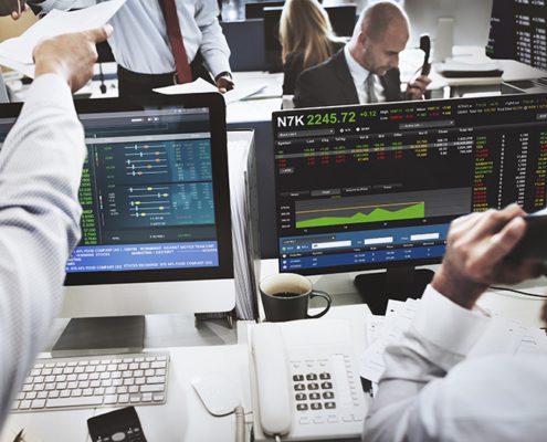 Several people working in a busy office with multiple computer screens displaying stock market data and analytics, communicating via phones and papers.
