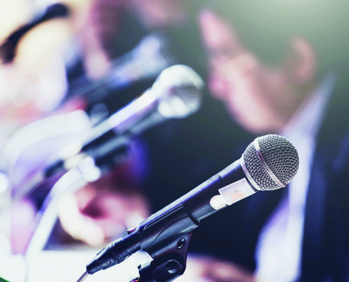 Close-up of a microphone on a stand, with a blurred person in the background, suggesting a conference or formal meeting setting.