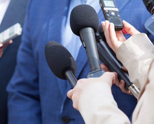 A person in a suit is surrounded by microphones and recording devices held by reporters, creating a press conference scene.