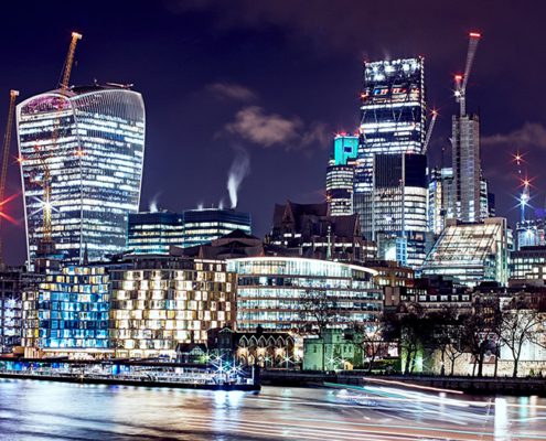 London cityscape at night featuring illuminated skyscrapers including The Gherkin, Walkie Talkie, and construction cranes along the Thames River.