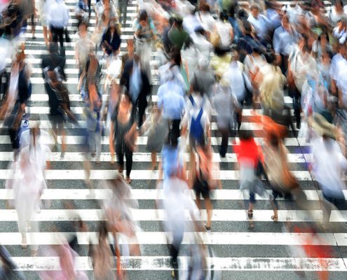 Blurred crowd of people crossing a busy striped crosswalk, implying motion and urban life, with no recognizable landmarks or historical buildings visible.