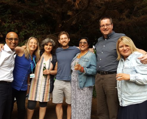 Seven people smiling outdoors, holding drinks, standing by trees. A table with bottles and glasses is partially visible in the foreground.