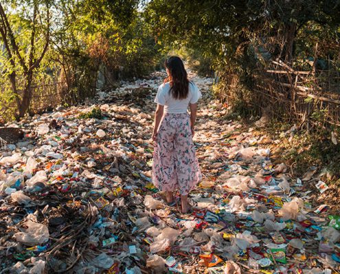 A person walks on a littered path surrounded by trees and scattered waste, capturing the impact of pollution on the environment.