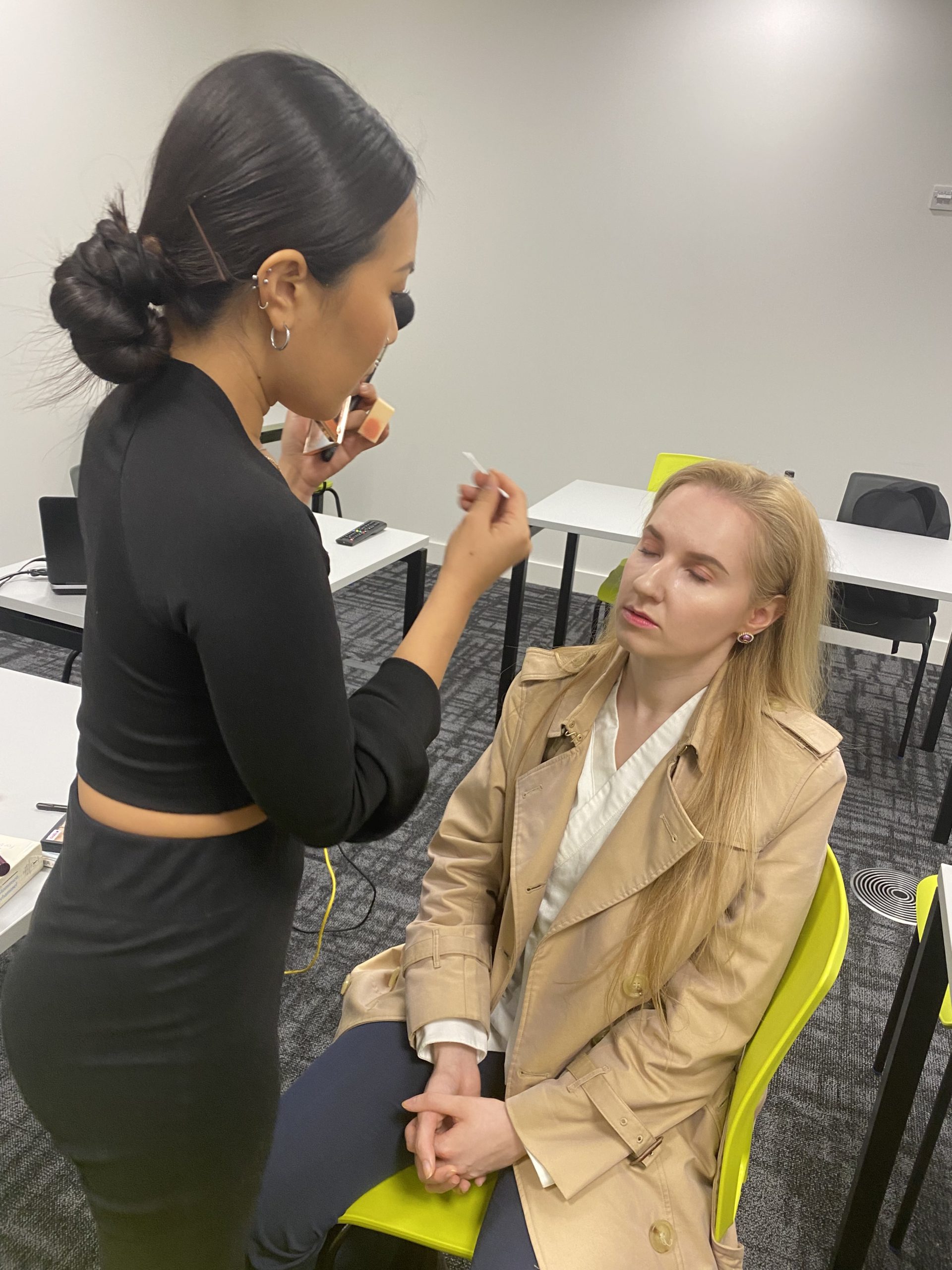A person applies makeup to another in a room with tables and chairs, suggesting a professional or educational setting.