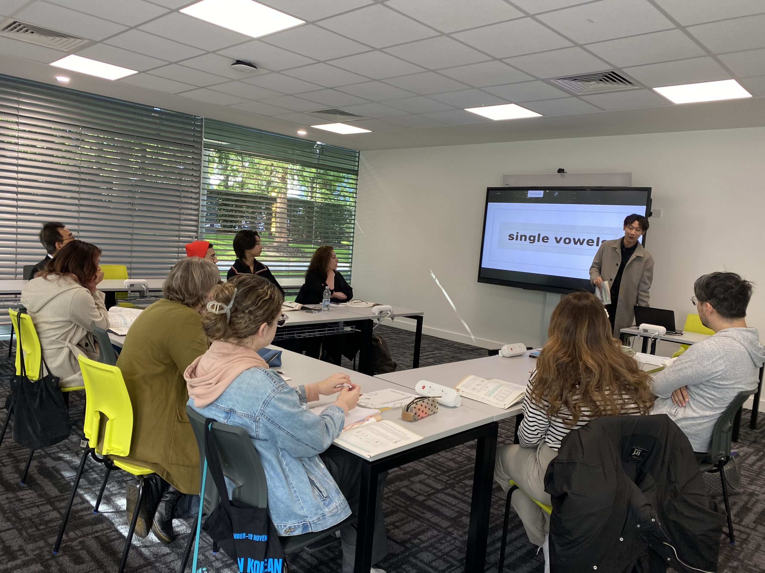 A group of people in a classroom setting, attentively listening to a presentation about "single vowels" displayed on a screen.