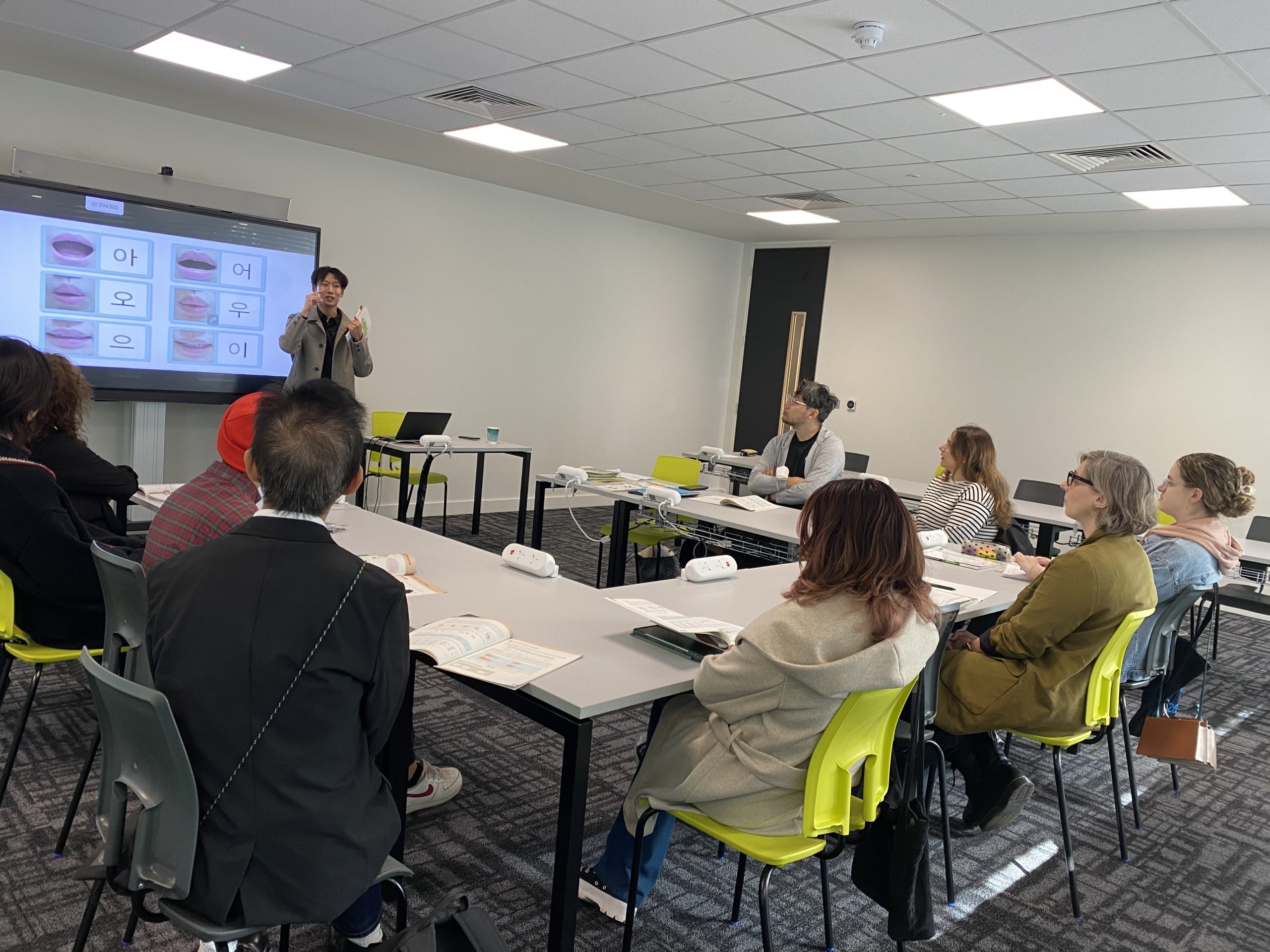 A person teaches a group in a classroom setting, using a screen displaying Korean characters and mouth positions for pronunciation practice.