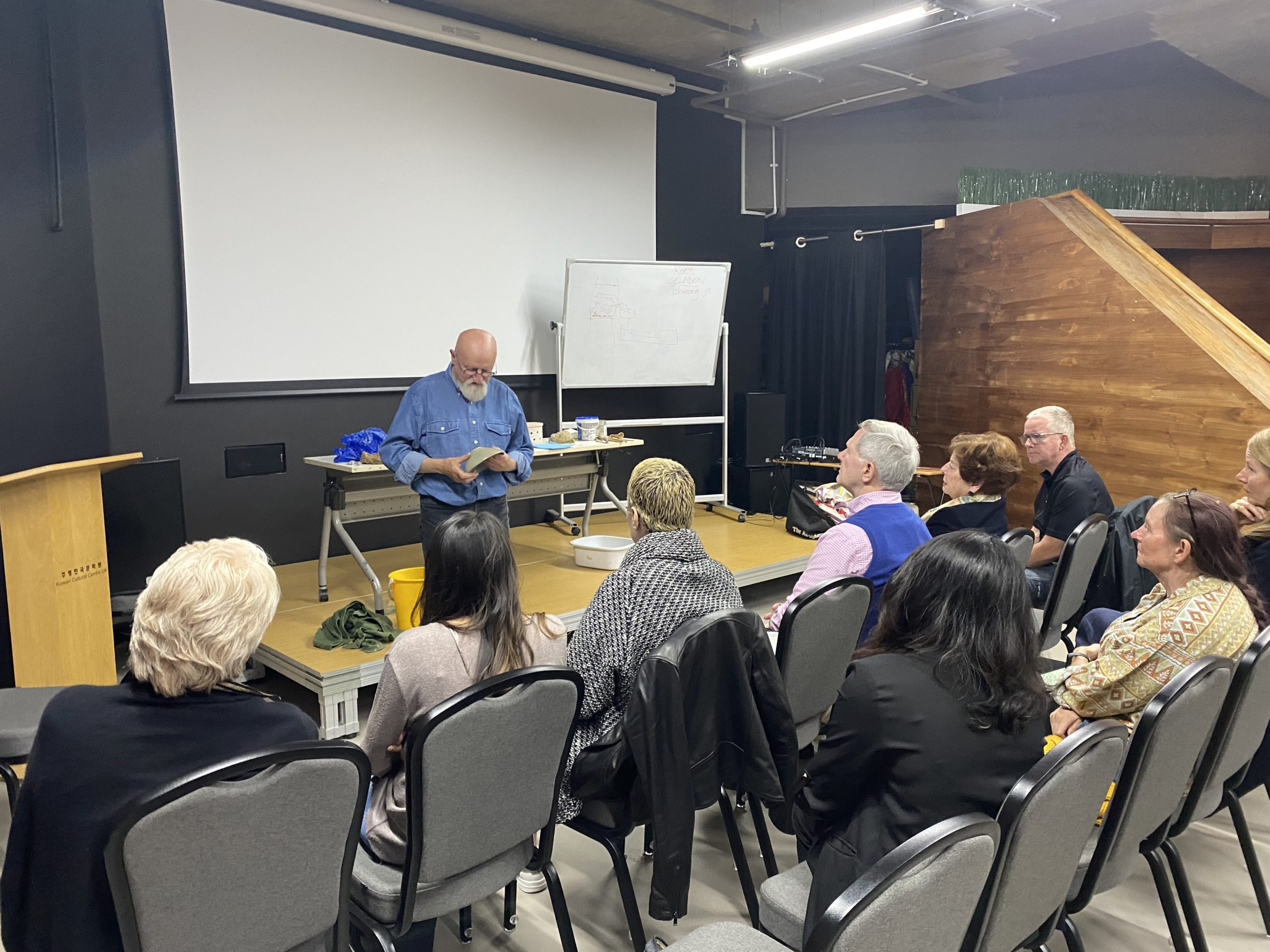 A person speaks to a group seated in a small lecture hall. A screen and whiteboard are in the background.