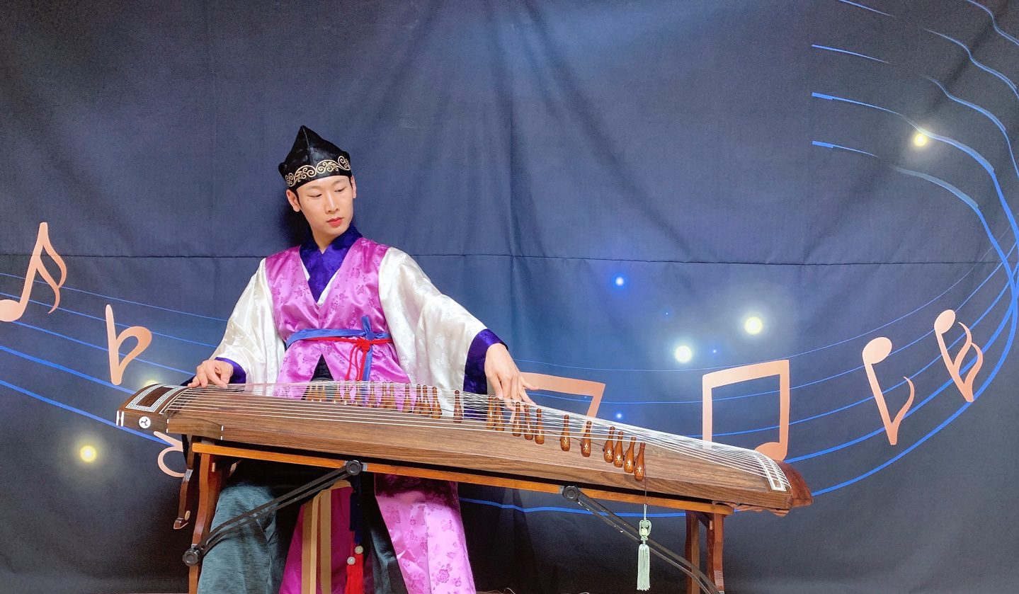 A person plays a traditional zither, wearing a colorful robe and hat. The background features musical notes and a dark backdrop.