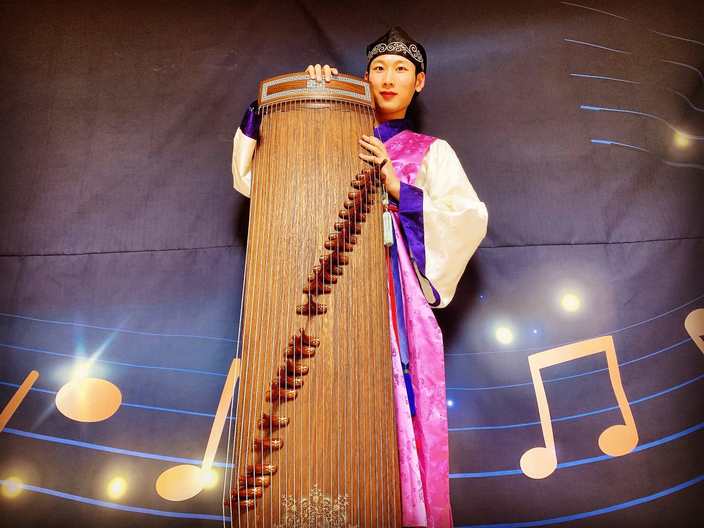 A person in traditional attire holds a large string instrument in front of a musical note-themed backdrop.