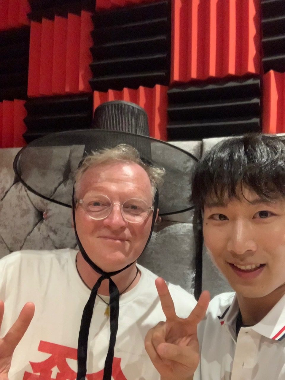 Two people pose with peace signs; one wears a traditional Korean gat hat. They're standing indoors against red and black acoustic panels.