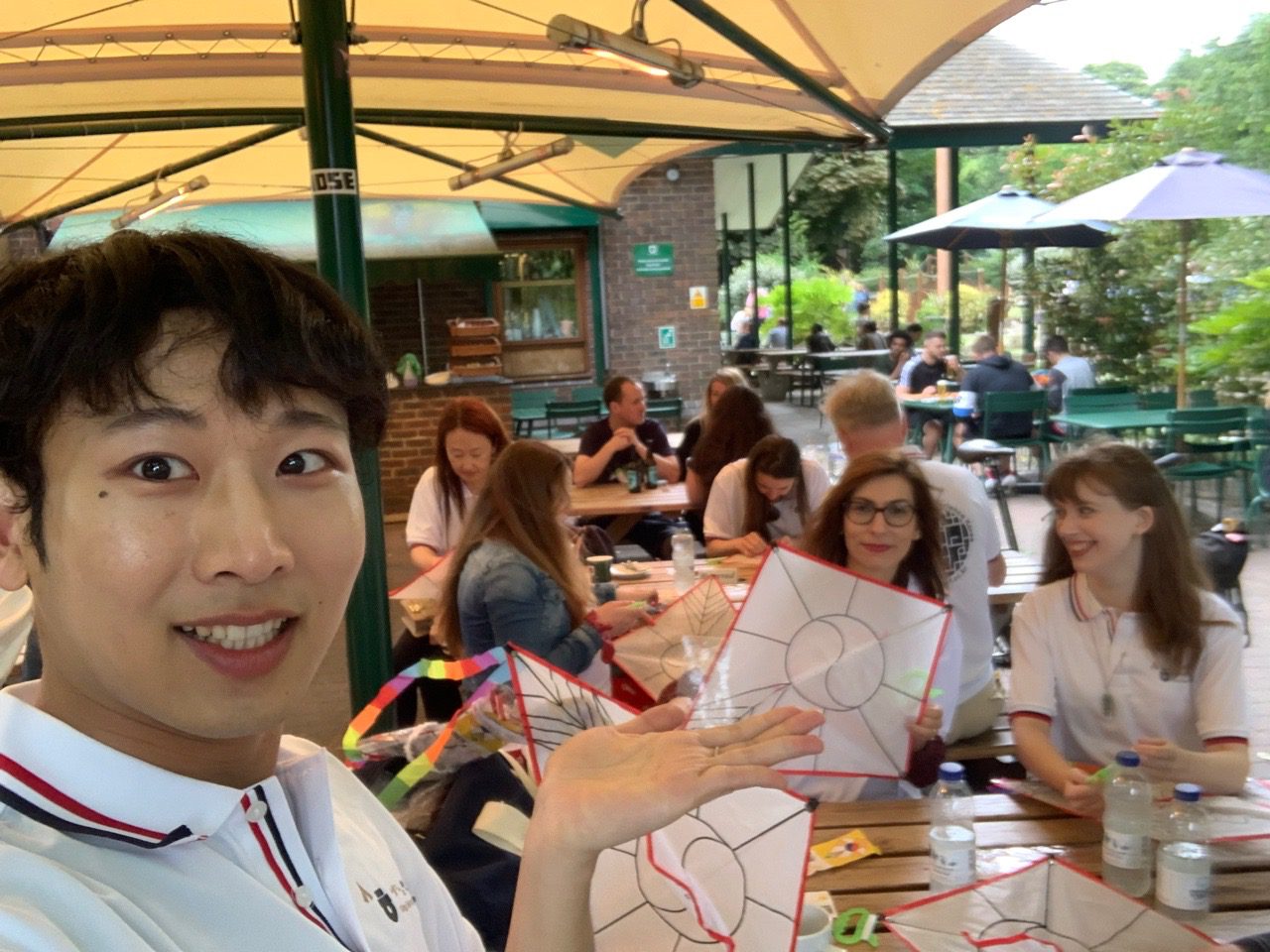 A person takes a selfie at an outdoor gathering. People in the background are socializing and engaging in creative activities at tables.
