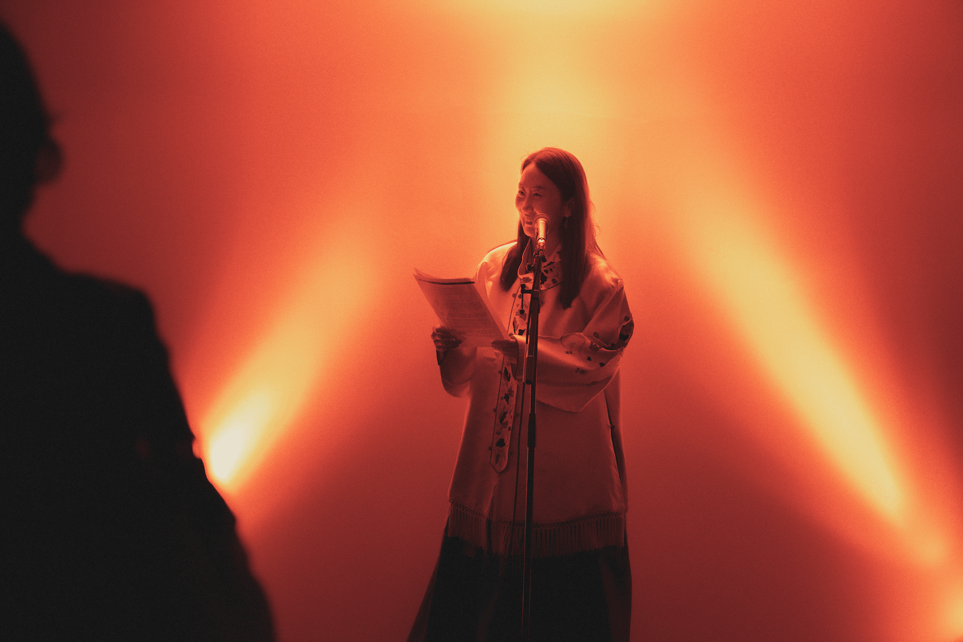 A person stands smiling under warm, orange lighting, holding papers and speaking into a microphone. Another person is silhouetted nearby.