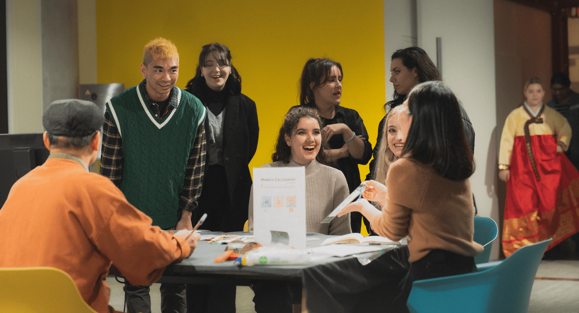 A group of people engaged in a lively workshop setting, with colorful backgrounds and materials labeled "Hangeul Calligraphy."