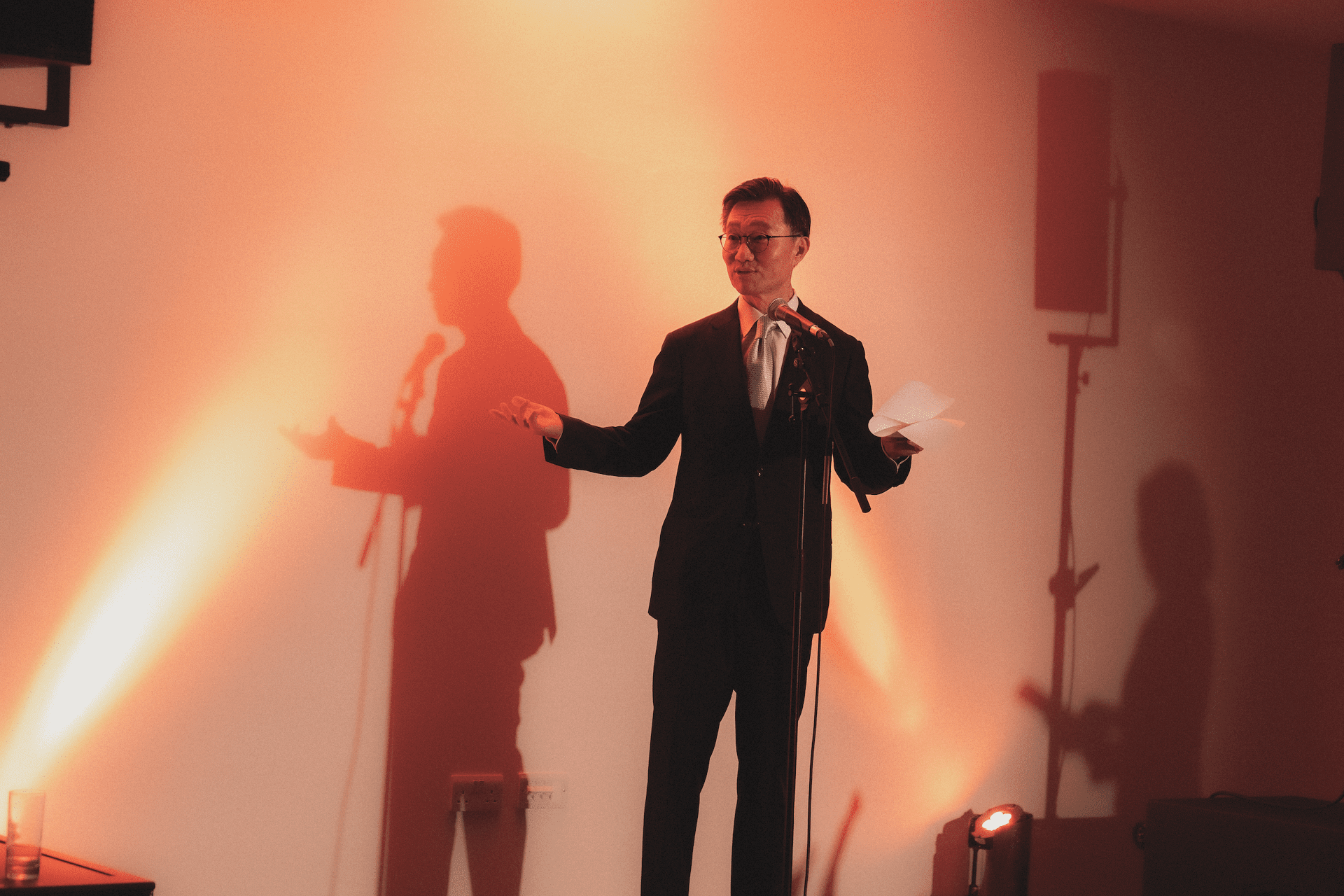 A person in a suit speaks into a microphone, casting a shadow on a warmly lit wall.