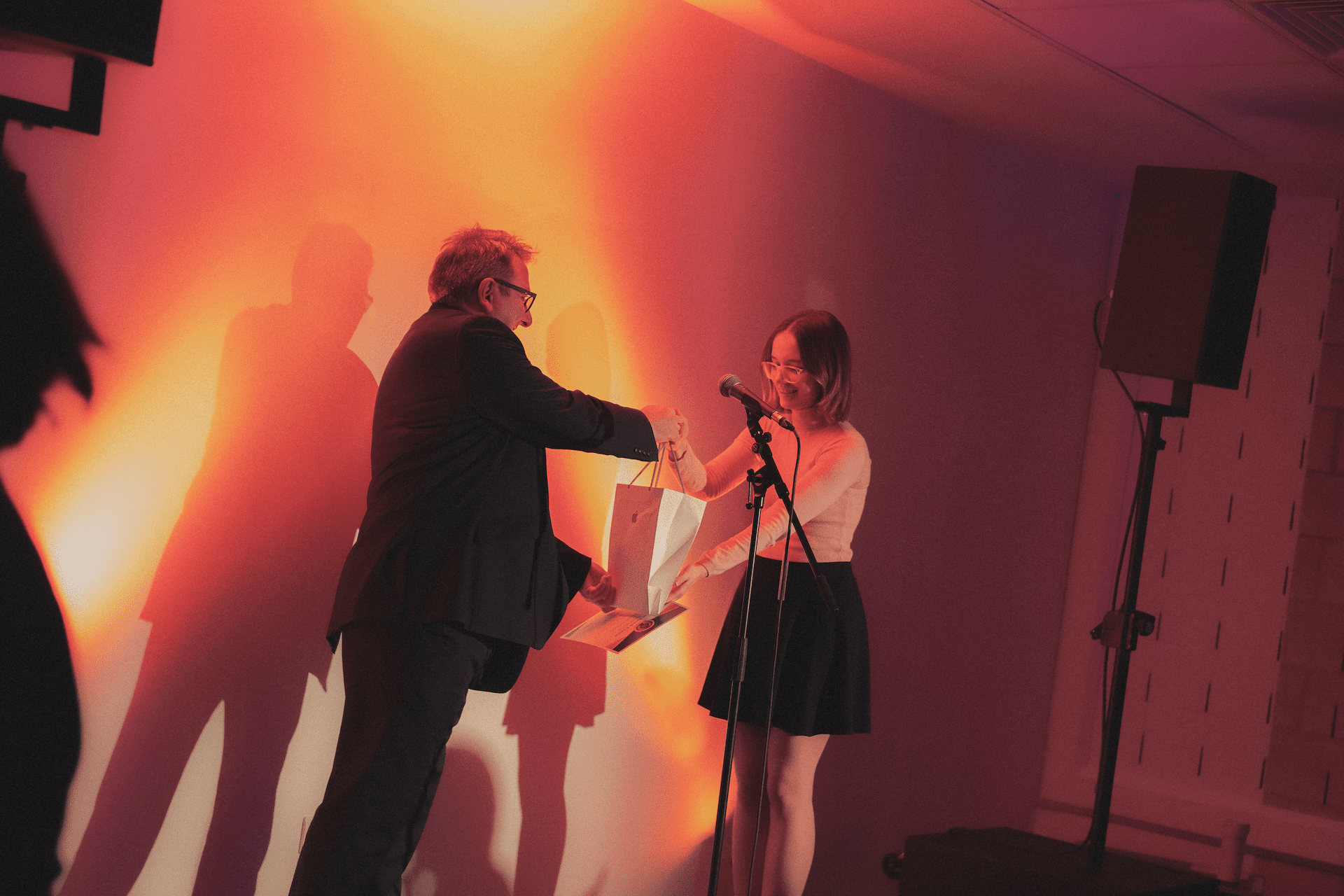 Two people exchange a gift on stage under warm lighting, with shadows cast on the wall behind them.